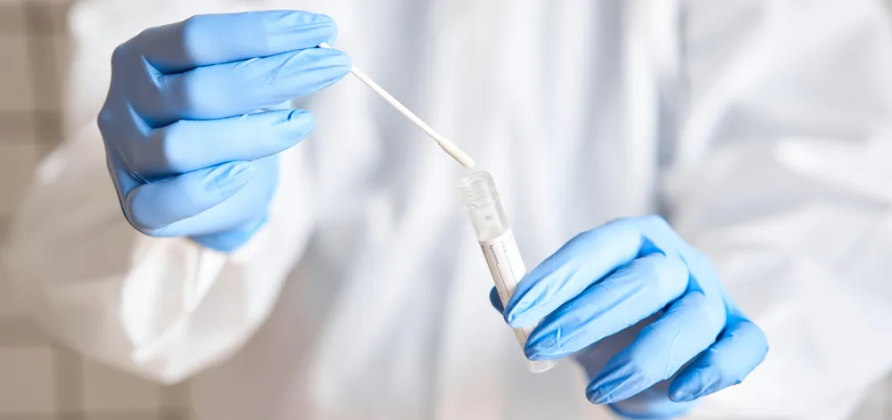 A-lab-tech-with-blue-latex-gloves-inserts-a-DNA-swab-into-a-test-tube-representing-the-fake-genomics-tests-that-Daniel-Hurt-ordered-to-defraud-Medicare
