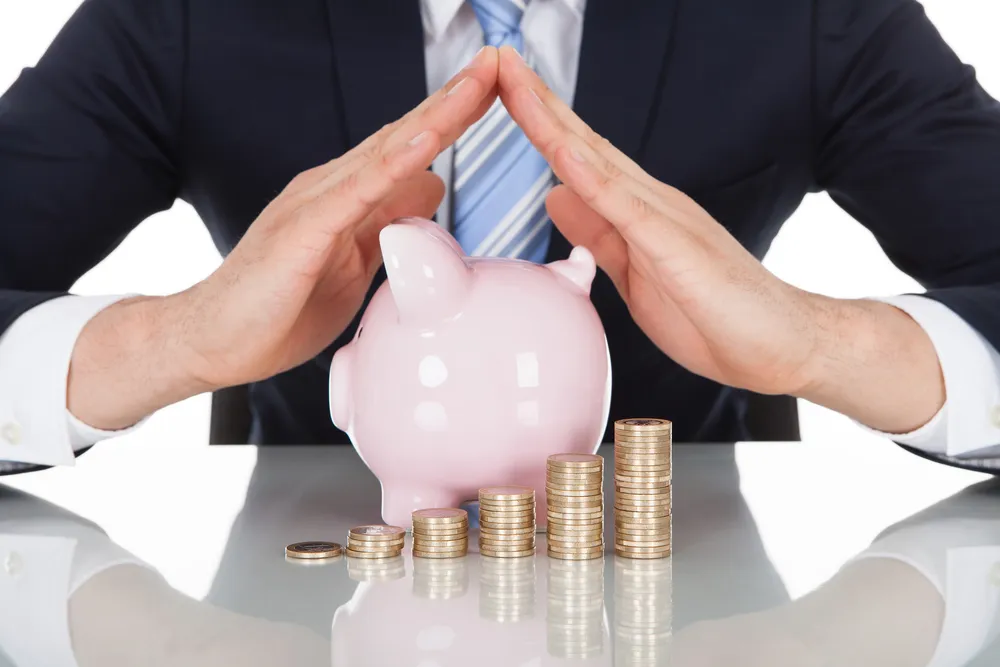 A Man Folds His Hands Over a Piggy Bank Besides Stacks of Coins as If Sheltering Them from Taxes As Stephen T. Mellinger III is Allegedly Guilty of Illegally Doing - The Daily Muck