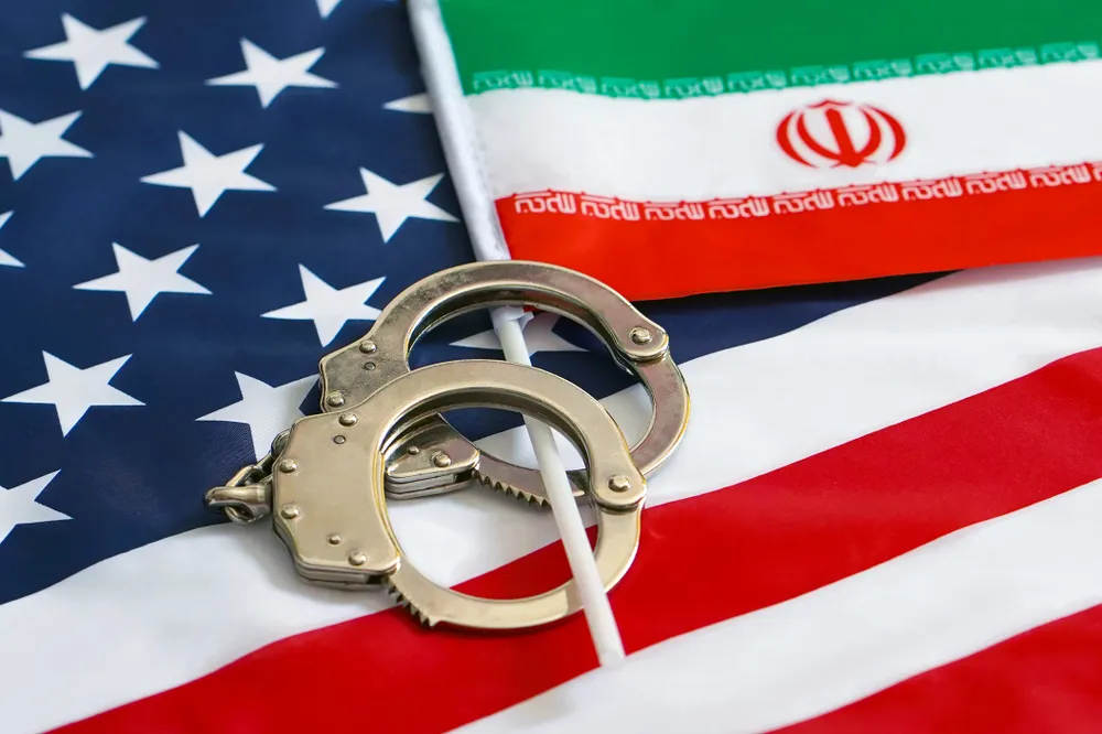 A Pair of Handcuffs Over an Iranian Flag on Top of An American Flag Represent Three Men Who Have Been Accused of Evading Iranian Sanctions to teh Detriment of the U.S - The Daily Muck
