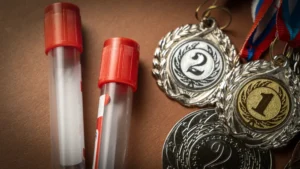 Two Vials of Blood Representing Doping Tests are Next to A Stack of Sports Medals Representing Doping Tests in Sports - The Daily Muck