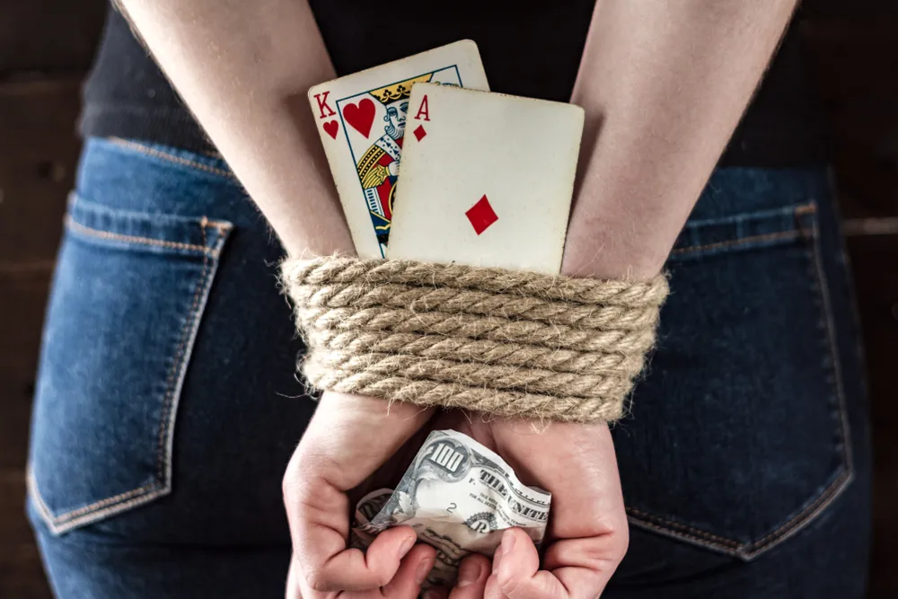 A Womans Hands are Tied with Rope Around Her Back and She is Holding Money and Playing Cards Representing Gambling Addiction. - The Daily Muck
