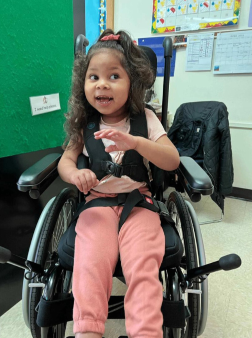 Six-year-old Ka'Mya Minor in a school wheelchair, smiling with her hand raised, wearing a pink outfit.