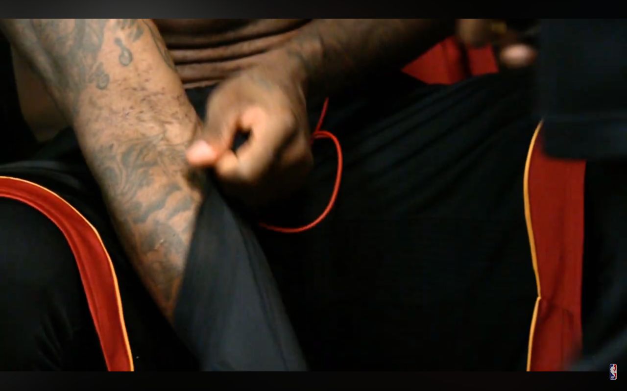 A close-up shot from an NBA Game 5 Mini-Movie showing LeBron James seated in a black and red Miami Heat uniform. As he uses his left hand to adjust the sleeve on his tattooed right arm, the lighting exposes his inner arm and the antecubital fossa (elbow crease). Visible on the skin are small, localized discoloration spots or puncture marks. These physical details are central to discussions regarding LeBron James steroids and PED allegations, serving as the visual evidence in debates over whether the scarring indicates routine IV hydration or track marks from performance-enhancing drug injections.